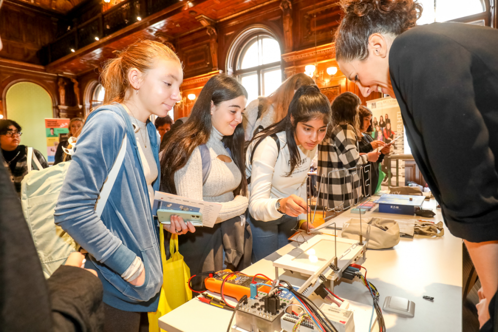 Drei Schülerinnen zeigen sich sich beim „Girls! Tech Up“ Erlebnistag bei einem Stand einer Dame sehr interessiert.