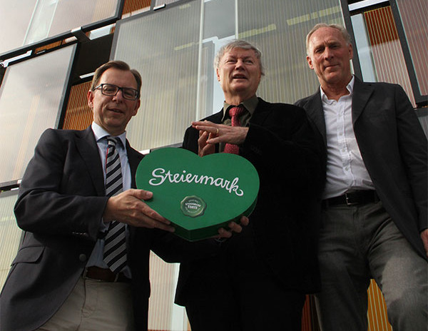 Three men standing in front of a building holding a heart-shaped Styria Cake.