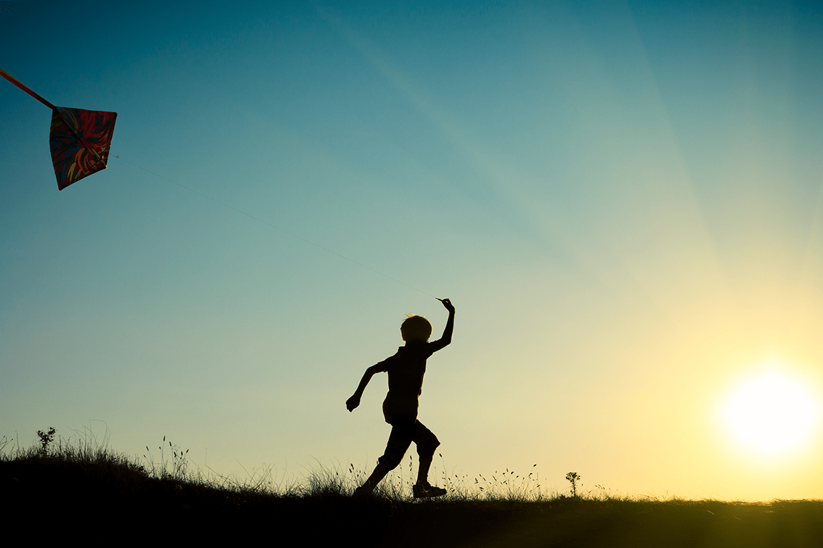 Boy flying a kite and running toward the sunset